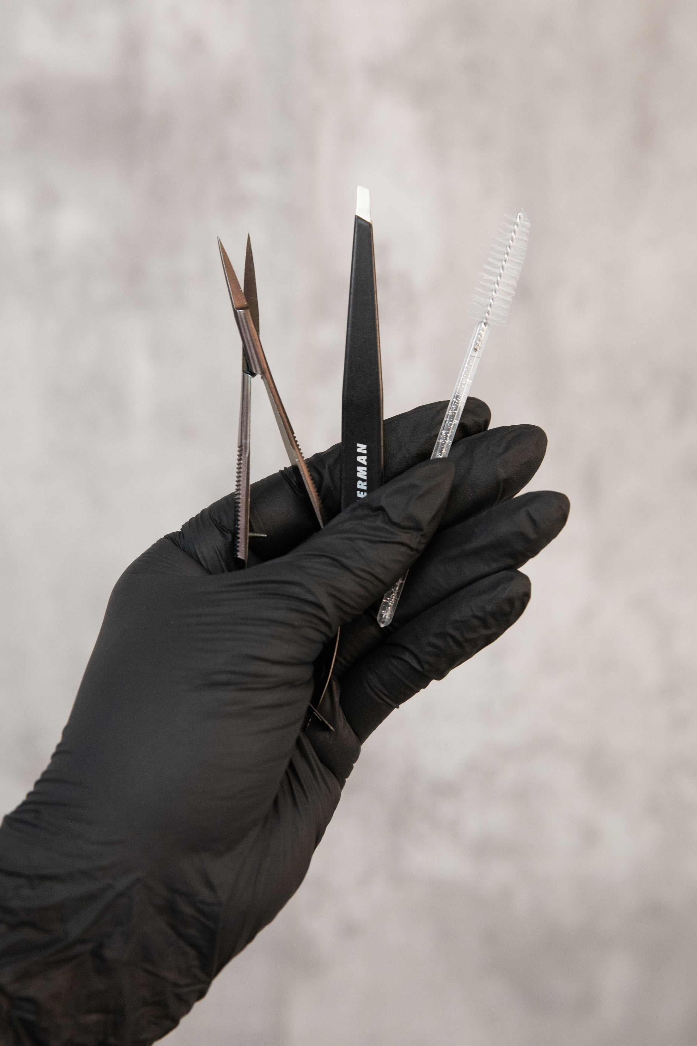 Hand wearing a black glove holding eyebrow waxing/ grooming tools against a neutral background